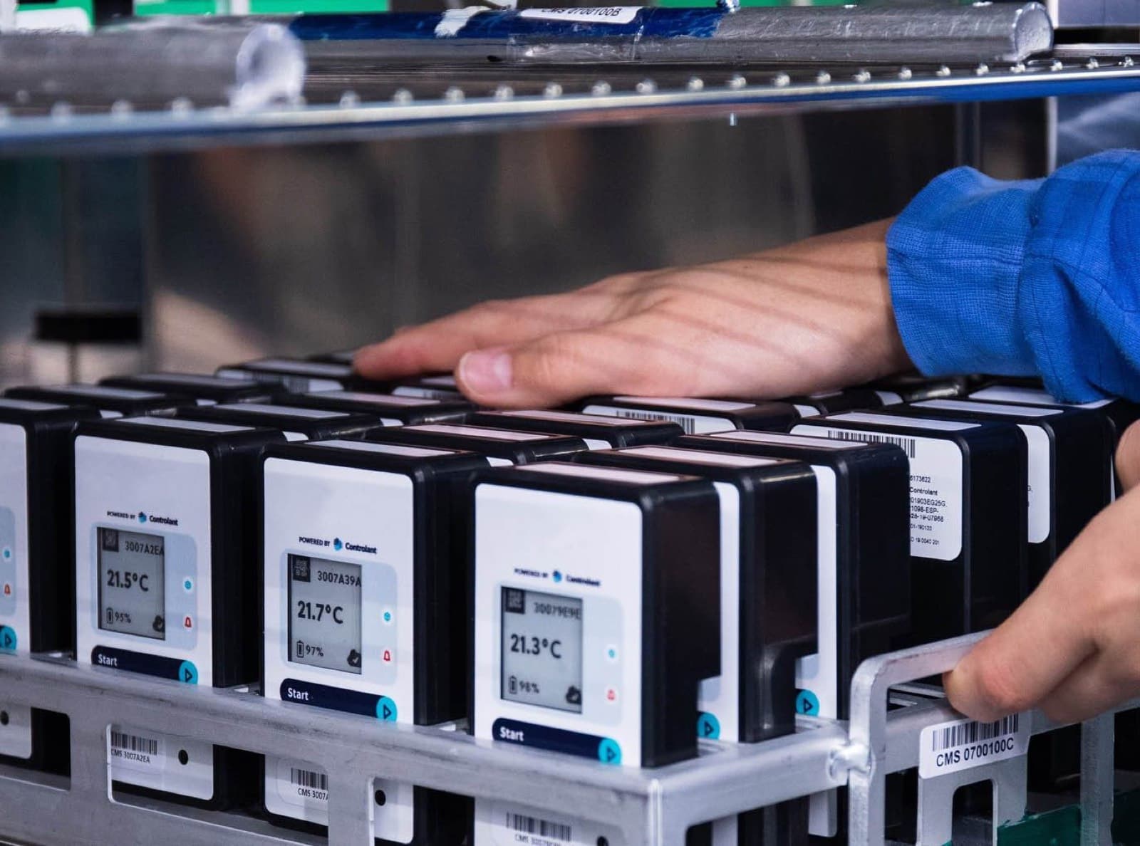 Hands at a metal rack of Saga devices, their screens reading 21.3 to 21.7 °C