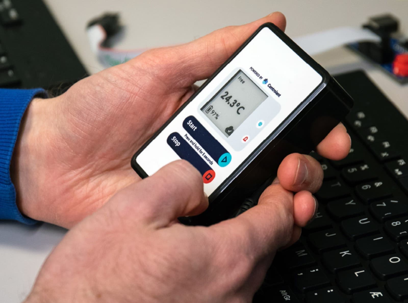 A hand holding a Controlant Saga device showing 24.3 °C, resting above a laptop keyboard